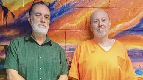 Rhys Wililams (l) - a white man with dark hair and a white beard and moustache stands alongside a balding man, also with a white beard and moustache, wearing an orange prison-issue top. The pair lean against a wall which has been painted and shows an orange, pink and purple sky.