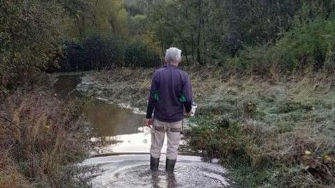 A man wearing wellies is standing in flood water which covers a path. The path runs through a field with grass, trees and bushes around it.