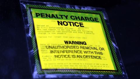 A yellow parking ticket is in a clear packet stuck to a windscreen and says Penalty Charge Notice