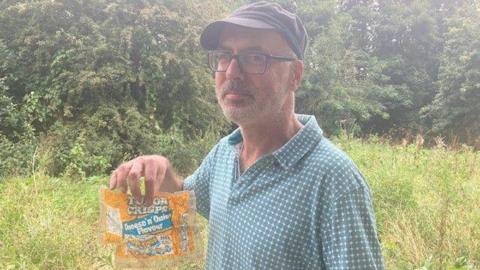 Steve Walsh, with a short grey beard and dark-rimmed glasses, holds an orange and blue Tudor Crisps packet.