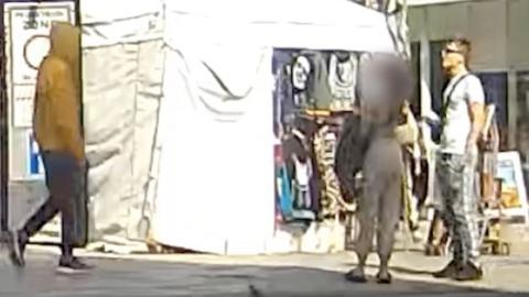 A boy in a yellow coat with the hood up approaches a man wearing grey T-shirt and tracksuit bottoms on a busy street.
