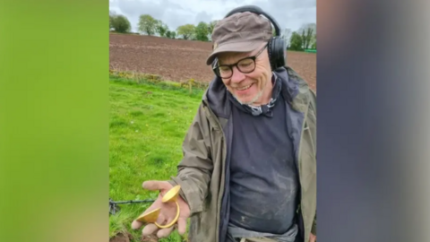 Johnathan is holding the item in his right hand as he stands in a field. He is wearing a scruffy jumper, jacket and cap. He is also wearing glasses. 