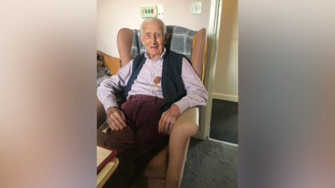 An elderly man is sitting in a chair in a care home, while wearing a fleece gilet and a pink shirt, as well as red wine coloured corduroy trousers. Attached to his chest is a medal.