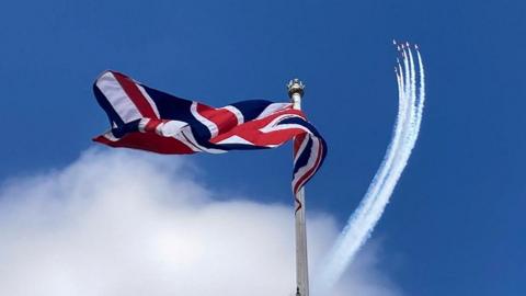 The Union Flag flies from a flagpole. In the distance, the Red Arrows aircraft in formation trail white smoke in a blue sky 