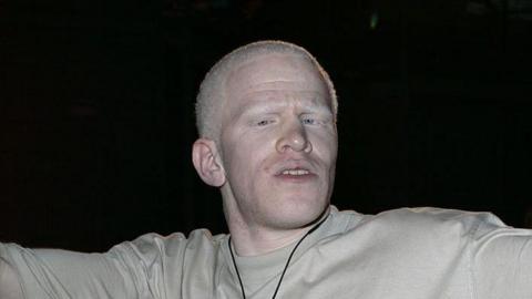 Darnell Swallow looks at the camera before he enters the Big Brother House. He wears a grey T-shirt and spreads his arms out from his body. He wears a microphone around his neck. 