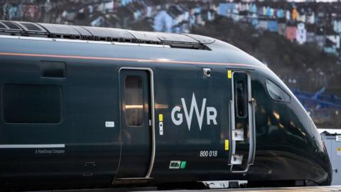 Image of a GWR train pulling into Bristol station. The dark green train takes up most of the image, with the Totterdown houses seen blurred in the background.