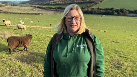 A blond woman with glasses and a green jacket in a large field with sheep behind her 