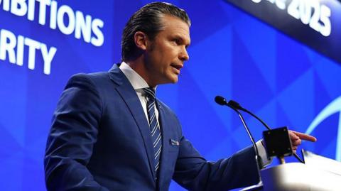 Pete Hegseth wearing a dark navy suit speaks at a lectern while pointing his finger at the audience. In the background is a blue screen displaying the Shangri-la Dialogue's name and topic of Hegseth's speech.
