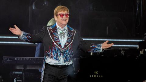 Elton John is standing on stage in a mostly dark jacket. He's wearing sunglasses with red lenses.