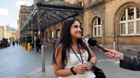 Girl stands in front of Glasgow Central train station speaking into a microphone.