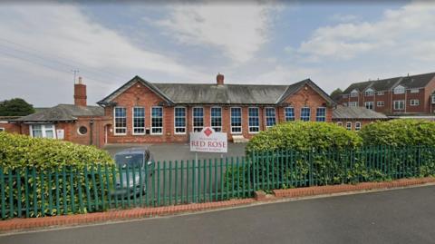 Green gates line a path. Bushes are behind them and a car park. A sign in the middle of the fence reads 'Welcome to Red Rose Primary School'. A red brick building sits behind the car park with large, long white windows. 