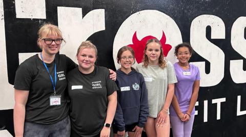 Five girls stand in front of a gym sign, they are wearing workout clothes and smiling.