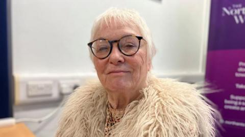 Danielle Stone smiles and looks directly at the camera. She is wearing a white, fluffy jacket and has tortoise shell-rimmed glasses on. She has short grey hair and is photographed indoors with a purple banner behind her.