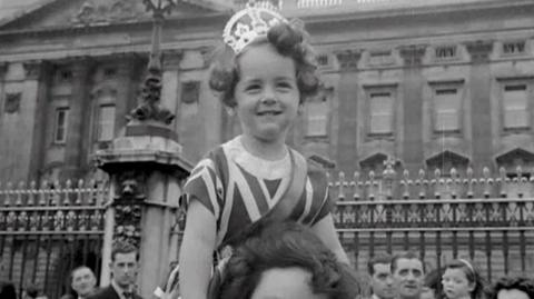 A girl wearing a crown and wearing a Union Jack dress outside of Buckingham Palace.