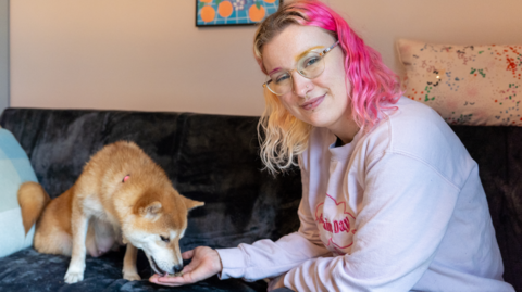 Ciara Pollen is smiling at the camera while Fern the dog eats treats from her outstretched hand. Ciara has blonde and pink wavy, shoulder-length hair and is wearing a pale pink sweatshirt. She and the dog are sitting on a sofa in a living room.