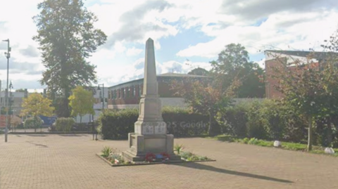 A small courtyard area with a war memorial in its centre. It is stone and tall with a square base that gets thinner towards the top. There are red poppy wreaths at its base, and small bits of greenery planted in a border around it.