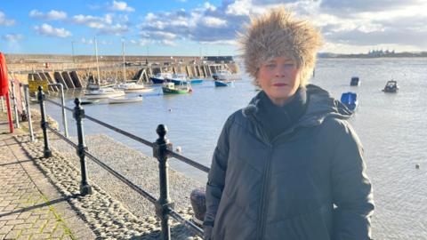 A woman in a coat and cossack-style hat stands in front of a harbour with some boats bobbing around. There is a harbour curving around on the left to her. The sky is blue, but features dark clouds.