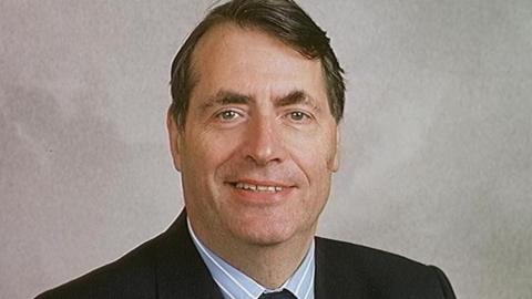 Sir Neville Trotter in a posed head and shoulders photo against a beige-grey background. He has short, dark brown and slightly receding hair. He wears a black suit jacket, striped blue and white shirt and a dark tie.