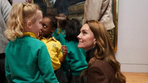 The Princess of Wales, wearing a brown jacket and black polo neck jumper, talks to one of the school children during their visit to the National Portrait Gallery