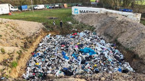 Picture shows a large amount of rubbish dumped in a three-sided pit. Two people can be seen in the background, along with several vehicles and containers.
