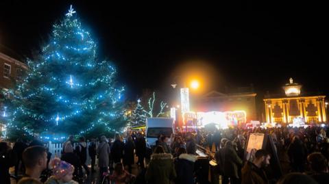 A night time Christmas light switch on in King's Lynn, showing a light Christmas tree, light up buildings, people standing bout, a white van, and street lights.