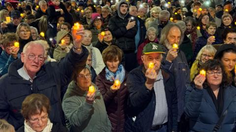 A large group of protesters hold candles up. They are standing close to each other. It's night. Everyone is wearing coats. 