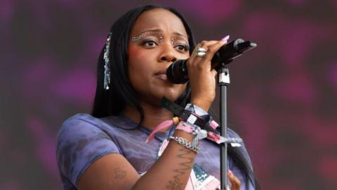 Rachel Chinouriri performs during day four of Glastonbury Festival 2024. Rachel is a woman in her 20s with straight black hair. She has brown eyes, made up with gems, and sings into a mic. She wears a purple top and festival wrist bands.