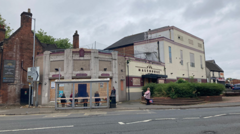 The view across a road to a derelict building with boarded up windows, with the words The Malthouse visible on the wall. The building varies in height, and is curved on the left hand corner where it meets the road
