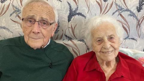 Tony and Joy sit side-by-side on a grey sofa, patterned with purple flowers. Tony is an older man with short white hair, and glasses. He wears a dark green jumper. Joy is an older woman, with short white hair. She wears a red blouse.
