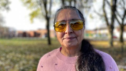 Woman in sunglasses with long hair and a pink jumper standing in front of trees.