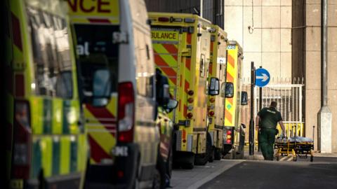 A row of ambulances queuing to get into a hospital. A paramedic in the distance is pushing an empty stretcher.