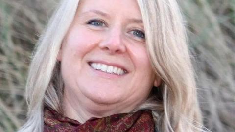 Close up image of Joanna Simpson's face. She is smiling and has light blond, medium-length hair. She is wearing a maroon scarf.