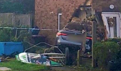 The side of a house with the rear end of a metallic blue car sticking out from a hole in the ground floor with the front door just beside it