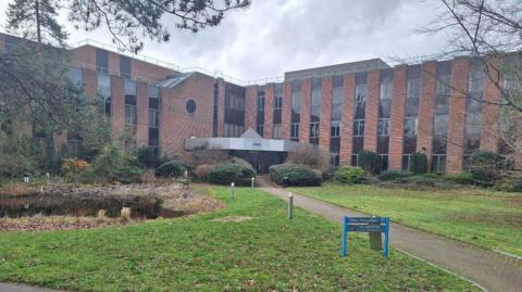 An office building with red bricks and black windows sits at the end of a footpath with grass on each side.
