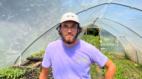 Man wearing purple t shirt stood in a polytunnel filled with growing plants.