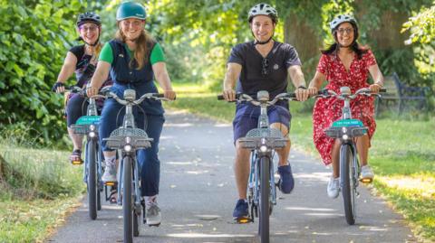 Four people wearing cycling helmets ride bikes along the path in the above picture.