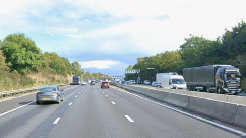 A motorway with a concrete central reservation barrier. There are trees on either side and a blue sky with white clouds in the background. Lorries, cars and vans are travelling along the road.