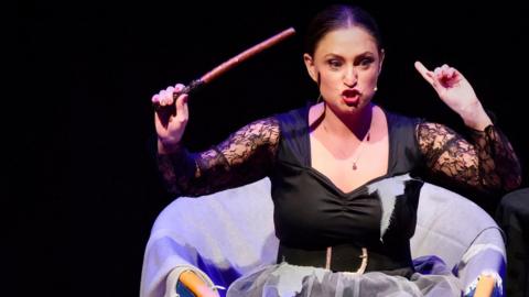 A woman dressed in a black witch's outfit, her black hair tied back, waves a wand and gestures expressively as she sits in an armchair covered with a pale lilac throw. The backdrop is black.