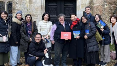 A group of eight Japanese tour operators and two representatives from Visit Britain pose with Caroline Lowsley-Williams, owner of Chavenage House, in the middle. Most are smiling at the camera and some are holding up a Chavenage House brochure. They are stood outside a large ornate black door, and the mottled Cotswold stone walls of the house can be seen behind them. 