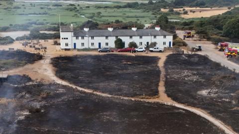 A large two-storey home surrounded by fields, the ones at the front scorched by fire. Fire engines can be seen parked to the right of the frame