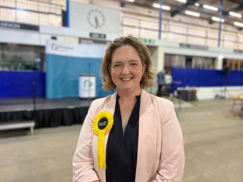 Anna Sabine standing with a pink jacket on. She has a black top on and a yellow rosette. She has brown/ blonde hair.
