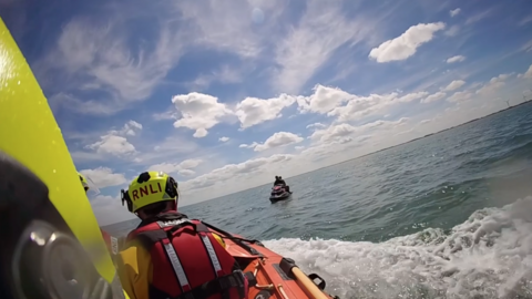 A shot from a body worn camera showing a crew member in a lifeboat in the sea. The person has their back to the camera and is dressed in a life jacket and a yellow helmet with "RNLI" written in red writing on the back. Two people on a water scooter can be seen nearby. The sky is blue and features white clouds.