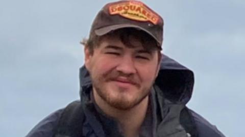 William is wearing a brown DSquared cap and navy blue Berghaus anorak. He is smiling at the camera.