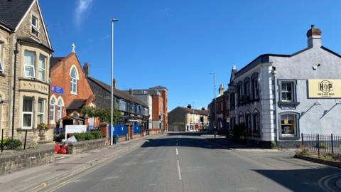Devizes Road, which is a two-laned highway with shops and housing on one side and a pub on the other side.