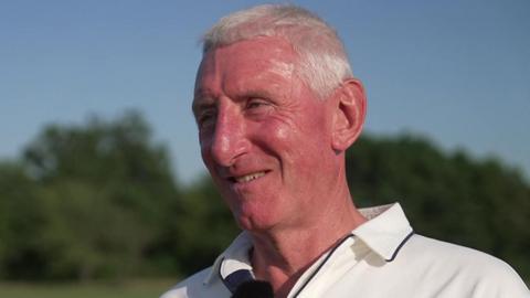 Nic Downes has short white hair and is smiling whilst looking off-camera. He is wearing cricket whites.