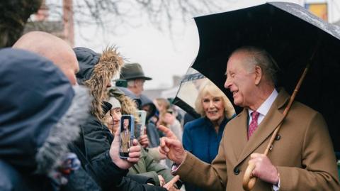 King Charles is in a smart brown double-breasted coat, with a red tie. He is holding a black umbrella over his shoulder. Queen Camilla is behind him in a blue coat. Charles is smiling or laughing while facing someone in the crowd. It is overcast above.