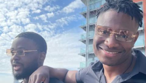 A close up of two people wearing matching orange-tinted sunglasses. The man on the right is smiling and has his arm on the shoulder of the man on the left, who looks happy but is looking away from the camera. Clouds are in the blue sky and a block of flats is in the background