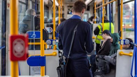 A conductor is stood on a Supertram with his back to the camera. He is wearing a navy blue uniform and carrying a bag and handheld ticket machine. Several passengers are sitting on the tram