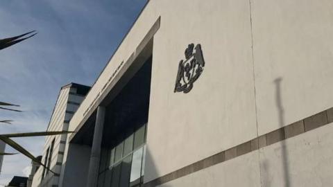 The white Isle of Man Courts of Justice building, which has the government crest on it,  with a blue sky above.