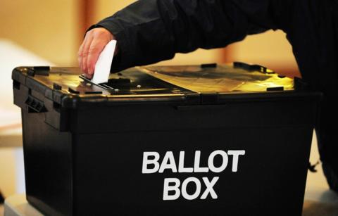 A hand drops a white ballot paper into a black box with the words ballot box written on the side in white